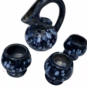 May include: A dark blue ceramic pitcher with a unique curved handle and spout, accompanied by three matching small cups. The set features a speckled white pattern, creating a contrasting design. The pitcher and cups are likely for serving beverages.
