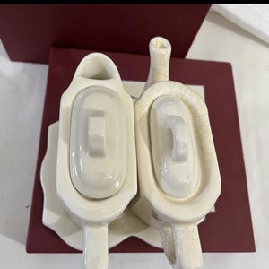 Puede incluir: Dos fuentes de cerámica blanquecina con tapas. Los platos tienen forma rectangular con un asa en la tapa. Los platos están sobre un plato blanco con fondo rojo.