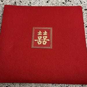 May include: A square, red invitation card with a gold-colored, decorative emblem in the center. The emblem features two Chinese characters, likely for a wedding or celebration. The card has a textured surface.