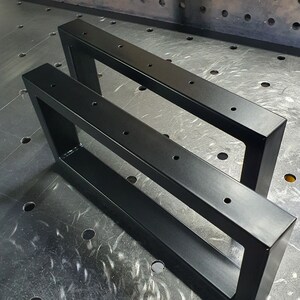 Op de afbeelding: Twee zwarte metalen tafelpoten met een rechthoekig frame en meerdere gaten. De poten staan op een metalen oppervlak met een patroon van gaten.