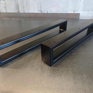 Op de afbeelding: Twee zwarte metalen tafelpoten met een rechthoekig ontwerp. Elke poot heeft een holle, open midden en voorgeboorde gaten voor montage. De poten zijn gemaakt van stevig metaal en hebben een matte afwerking.