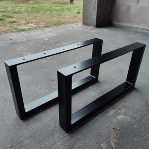 Op de afbeelding: Twee zwarte metalen tafelpoten, waarvan er één iets groter is dan de andere, zijn te zien op een grijs betonnen oppervlak. De poten zijn rechthoekig en hebben een eenvoudig, modern ontwerp.