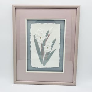 May include: Framed artwork featuring white daffodils with yellow centers and green leaves. The daffodils are set against a textured white background with a blue-gray border, all within a pinkish-gray frame. A botanical-themed piece.