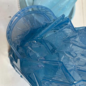 May include: Close-up of a blue plastic toy castle with glitter accents. The toy has a multi-faceted design and appears to be part of a larger playset. The color is a translucent blue, with a light blue castle structure in the background.