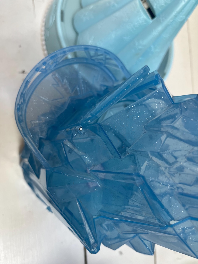 May include: Close-up of a blue plastic toy castle with glitter accents. The toy has a multi-faceted design and appears to be part of a larger playset. The color is a translucent blue, with a light blue castle structure in the background.