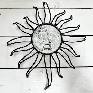 Puede incluir: Decoración de pared de sol de metal negro con una cara de cristal. El sol tiene largos rayos curvos que se extienden hacia afuera desde una cara circular central. La cara tiene un diseño grabado detallado. Decoración sobre superficie de madera blanca.