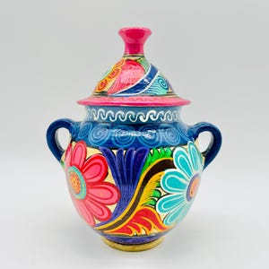 May include: A hand-painted ceramic jar with a lid, featuring two side handles and a vibrant floral pattern. The design includes large pink and turquoise flowers, yellow and red leaves, and blue swirls on a dark blue background with a pink lid knob.