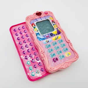 Puede incluir: Teléfono de juguete rosa con teclado y pantalla. El teléfono presenta imágenes de princesas de Disney y un botón en forma de corazón. El teclado tiene letras y gráficos de princesas. El teléfono tiene un teclado numérico.