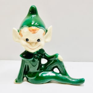 Puede incluir: Una figurita de elfo de cerámica verde con la cara blanca y un gorro rojo y naranja. El elfo está sentado con las piernas cruzadas y los brazos cruzados.