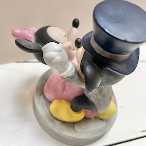 May include: A porcelain figurine of Mickey and Minnie Mouse in an embrace. Mickey wears a black tuxedo and top hat, while Minnie dons a pink dress and bow. The base is a gray, circular design.