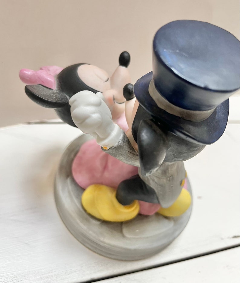 May include: A porcelain figurine of Mickey and Minnie Mouse in an embrace. Mickey wears a black tuxedo and top hat, while Minnie dons a pink dress and bow. The base is a gray, circular design.