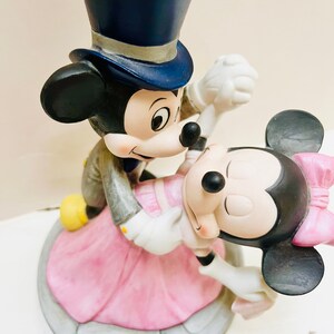 May include: A ceramic figurine of Mickey Mouse and Minnie Mouse dancing. Mickey wears a blue top hat and gray suit, while Minnie dons a pink dress and bow. The base is a light gray color.