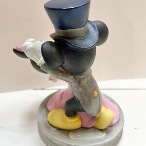 May include: A ceramic figurine of Mickey Mouse in a tuxedo and top hat, holding Minnie Mouse's hand. Minnie is wearing a pink dress. The base is gray with a circular design. Mickey's shoes are yellow.