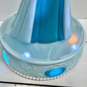 May include: A light blue Elsa-themed night light. The base has a decorative design with the name "Elsa" on the front. The night light has a light blue and white dress design with snowflake details. The base has several small, glowing lights.
