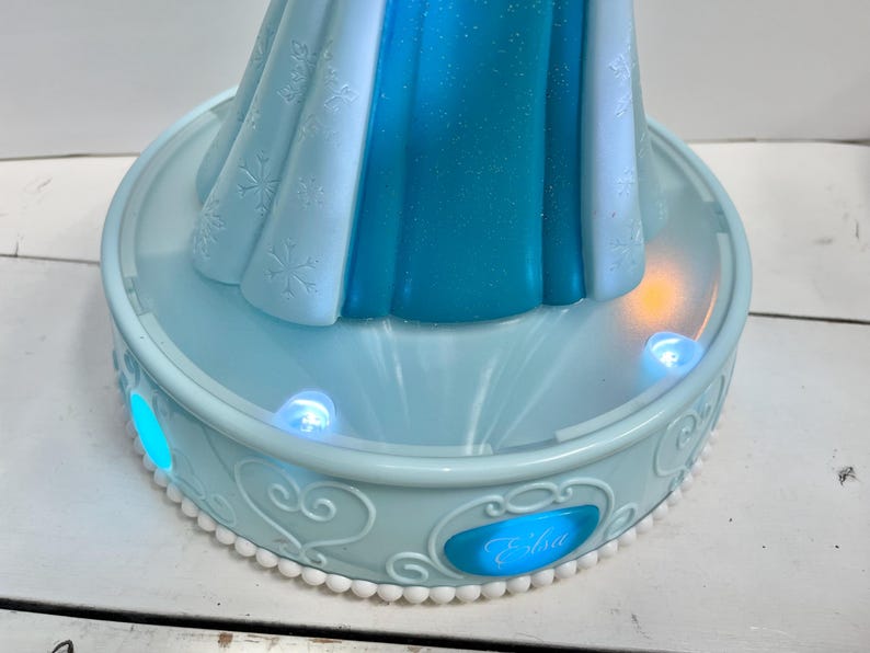 May include: A light blue Elsa-themed night light. The base has a decorative design with the name "Elsa" on the front. The night light has a light blue and white dress design with snowflake details. The base has several small, glowing lights.