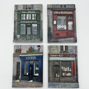 May include: Four decorative rectangular wall plaques depicting French storefronts. One reads "Chez Jerome Restaurant 62", another "Vins de Bourgogne", a third "Livres", and the last "France-Soir Librairie Papeterie". Each plaque features detailed architectural elements.