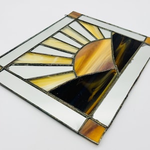 May include: Stained glass suncatcher with a geometric design. The sunburst features yellow, orange, and black glass, framed by mirrored glass and brown accents. A small metal loop is attached for hanging.
