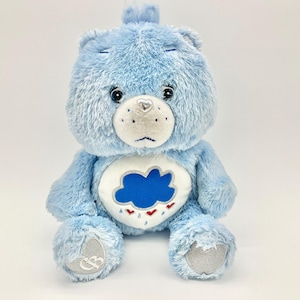 Puede incluir: Un oso de peluche azul claro con un hocico blanco y un corazón plateado en la nariz. El oso tiene un diseño de nube y lluvia en el vientre. Es un peluche.