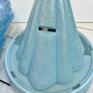 May include: A light blue plastic toy shaped like a dress, with snowflake patterns embossed on the surface. The toy sits on a matching light blue circular base with a decorative edge. A slot is visible on the dress.