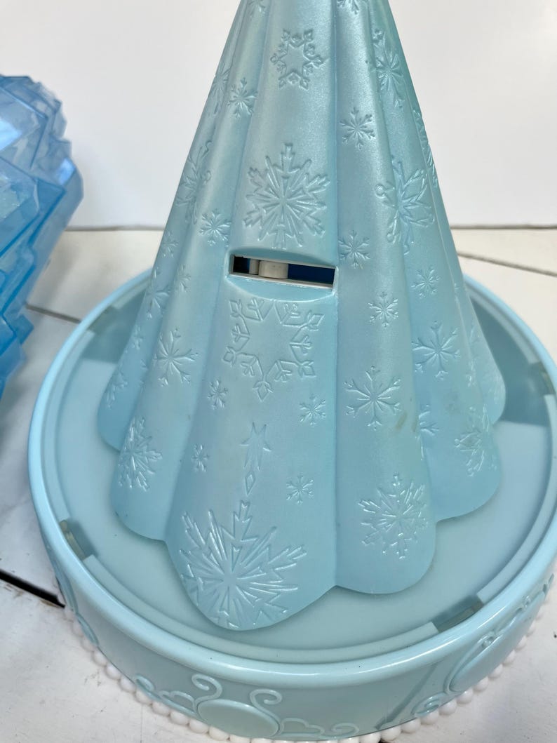 May include: A light blue plastic toy shaped like a dress, with snowflake patterns embossed on the surface. The toy sits on a matching light blue circular base with a decorative edge. A slot is visible on the dress.