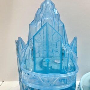 May include: A light blue plastic castle toy with a detailed design. The castle features a multi-tiered structure with snowflake patterns and a balcony. The toy is designed to resemble an ice castle.
