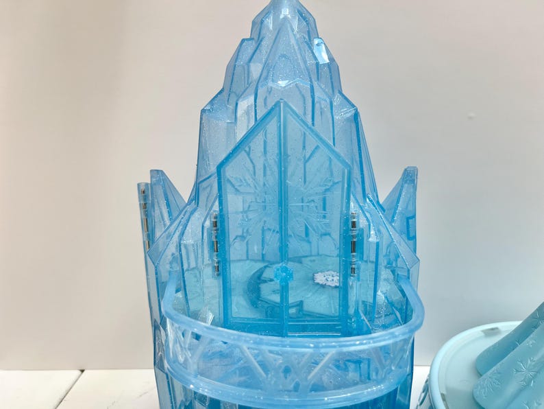 May include: A light blue plastic castle toy with a detailed design. The castle features a multi-tiered structure with snowflake patterns and a balcony. The toy is designed to resemble an ice castle.