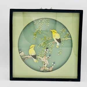 May include: A framed artwork featuring two yellow birds perched on branches with green leaves and white flowers. The birds have black and white markings. The artwork is set against a light green background within a black frame.