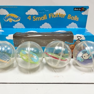 May include: Four clear plastic balls with Teletubbies characters inside. The balls are labelled "4 Small Flutter Balls" and "Born to Play" on the box.