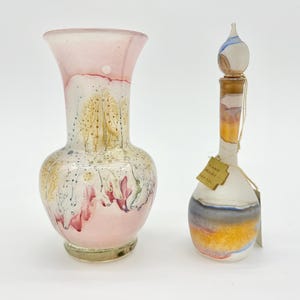 Puede incluir: Un jarrón y una botella decorativos de cristal. El jarrón es rosa y blanco con detalles florales. La botella tiene un diseño colorido con un corcho y una etiqueta "Hand Made".