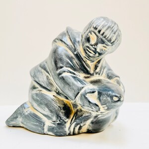 May include: A ceramic sculpture of a person in a hooded garment holding a smaller figure. The sculpture is painted in shades of gray and white.