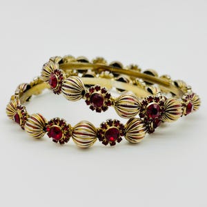 Peut inclure: Deux bracelets dorés avec des accents de pierres précieuses rouges. Les bracelets présentent un motif répétitif de perles côtelées et de montures en forme de fleurs, chacune contenant une pierre précieuse rouge ronde. Les bijoux sont sur fond blanc.