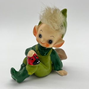 Puede incluir: Figura de cerámica vintage de un elfo sentado, con un atuendo verde, gorro puntiagudo y un mechón de pelo claro. El elfo sostiene una mariquita roja con manchas negras. La figura tiene una expresión alegre y es un objeto de colección.