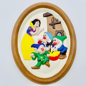 May include: Oval wall plaque depicting Snow White and the Seven Dwarfs in relief. Snow White wears a yellow dress, and the dwarfs are in colourful outfits. The plaque has a brown wooden frame and a cream background.