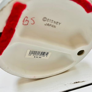 May include: A white ceramic figurine base with the text "&copy; DISNEY JAPAN". A sticker with a barcode and numbers is attached. Red tape and handwritten text are also visible.