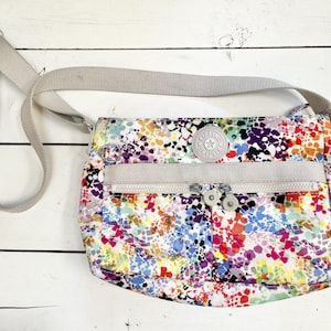 May include: A colorful crossbody bag with a white background and a multi-colored abstract pattern. The bag has a beige strap and a zipper closure. The bag has a small round logo with the text "Kipling" on it.