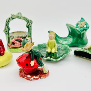 May include: A collection of vintage ceramic figurines featuring elves and fairy-tale elements. The figurines include a red-clad elf on a yellow crescent, a green archway, a green shoe, and elves on leaves and a ladybug.