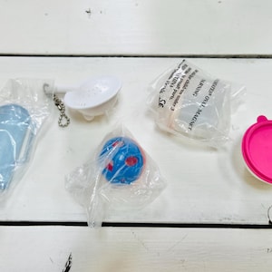 May include: A collection of five small items, including a blue and red ball, a white strainer with a handle, a pink lidded container, a blue container, and a clear plastic container with the text "LITTLE OVAL MAGNET 3 magnets under 3 CE WARNING".