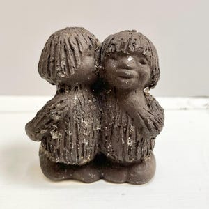 May include: A dark brown ceramic sculpture of two figures embracing. The figures have textured, shaggy hair and bodies, with a simple, rounded base. The sculpture has a rustic, handmade appearance.
