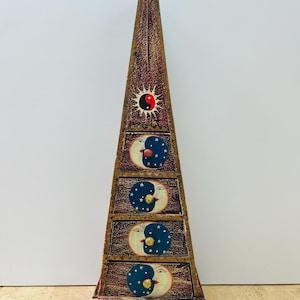 May include: A pyramid-shaped wooden cabinet with four drawers, decorated with celestial and spiritual designs. The cabinet is brown with gold detailing. Each drawer has a crescent moon and star design. The top features a sun and yin-yang symbol.