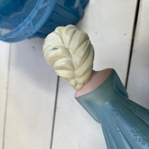 May include: A light blue plastic figurine of a character with a blonde braid, wearing a blue dress with snowflake patterns. The figurine is part of a larger toy set, with a blue plastic dome visible in the background.