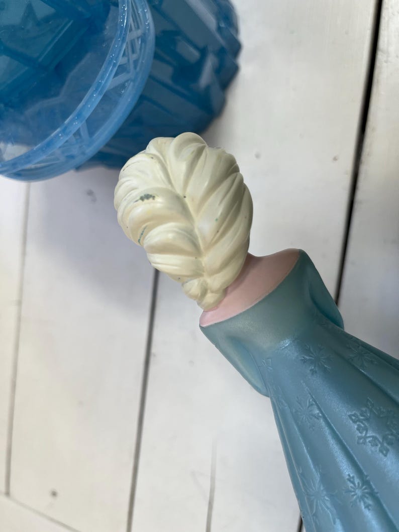 May include: A light blue plastic figurine of a character with a blonde braid, wearing a blue dress with snowflake patterns. The figurine is part of a larger toy set, with a blue plastic dome visible in the background.