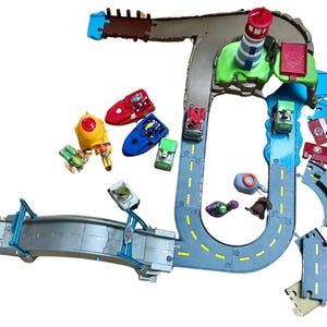 May include: A colorful toy playset featuring a road track, a lighthouse, and various vehicles. The set includes a red and yellow launch pad, a gray bridge, and several small toy cars and boats. The track is gray with yellow lane markings.