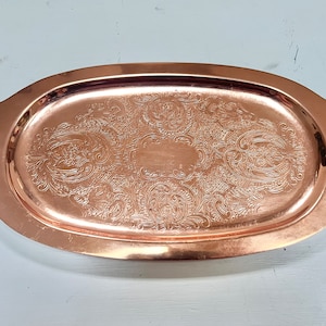 May include: An oval copper serving tray with an ornate, embossed design. The tray has a slightly raised edge and a smooth, polished finish.