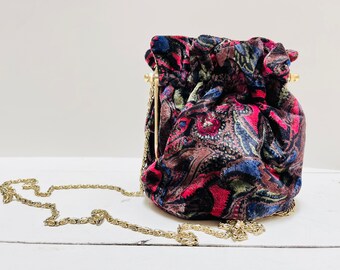 May include: A small, round, drawstring bag with a gold chain strap. The bag is made of a dark fabric with a paisley pattern in shades of pink, blue, and green.