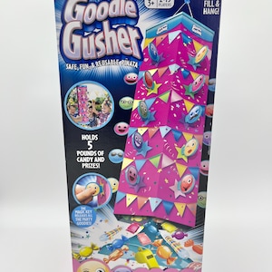 Puede incluir: Una caja de juego Goodie Gusher. La caja es azul y rosa con las palabras "Goodie Gusher" en letras grandes. La caja dice que contiene 2,27 kg de caramelos y premios. El juego es para niños de 3 años o más y de 2 a 15 jugadores.