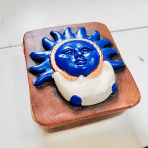 以下が含まれることがあります： 青と白の太陽と月のデザインが施された木製の箱。太陽は人間の顔をしていて、光線が放射状に広がっています。月は三日月形です。