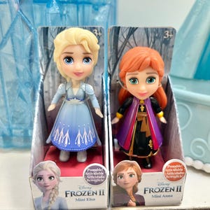 May include: Two Frozen II Mini dolls, Elsa and Anna, in their original packaging. Elsa wears a blue dress, and Anna wears a black and purple outfit. The packaging features character images and the text "Frozen II".