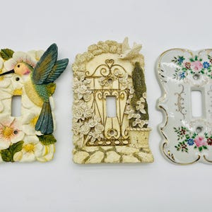 Puede incluir: Tres cubiertas decorativas para interruptores de luz. Una presenta un colibrí y flores, otra una puerta de jardín con rosas y la tercera un diseño floral con ribetes dorados. Todas tienen una abertura rectangular.