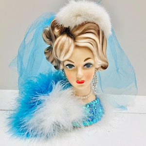 Puede incluir: Una cabeza de cerámica vintage con cabello rubio, labios rojos y ojos azules, adornada con un sombrero de plumas blanco, tul azul y una parte superior con lentejuelas azules. La cabeza también presenta aretes colgantes de diamantes de imitación y un collar de perlas.
