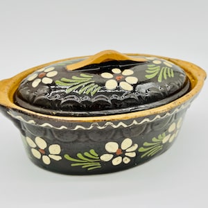 以下が含まれることがあります： 楕円形の黒いセラミックキャセロール皿と蓋。皿と蓋は、白とオレンジの花と緑の葉で飾られています。縁とハンドルはタン色です。